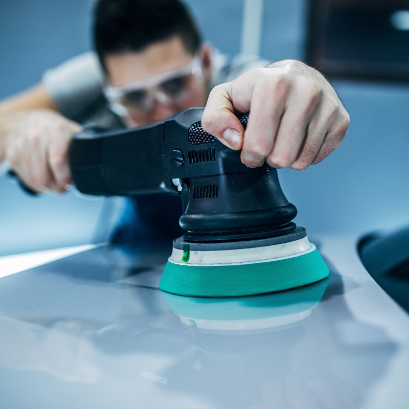 Un homme aux lunettes de protection polit la carrosserie grise d'une voiture avec une lustreuse électrique à pad vert, pour une brillance éclatante.