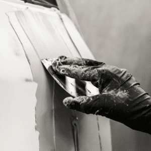 Main gantée appliquant du mastic à la spatule sur une carrosserie de voiture en réparation. Travail de préparation de surface.